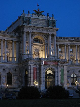 Das Bild zeigt die nächtlich beleuchtete Fassade der Österreichischen Nationalbibliothek in Wien. Das imposante Gebäude im barocken Stil ist detailreich dargestellt, mit seinen Säulen, Statuen und dem großen Eingangsportal. Ein Banner mit der Aufschrift "Österreichische Nationalbibliothek" ist sichtbar. Der dunkle Himmel bildet einen starken Kontrast zur hellen Beleuchtung des Gebäudes.