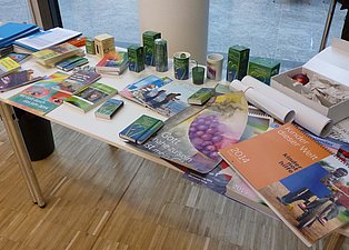 Das Bild zeigt einen Tisch mit verschiedenen Artikeln darauf, darunter Bücher, Broschüren, Kalender, Kerzen, Streichhölzer und andere kleine Gegenstände. Die Artikel hängen thematisch zusammen. Der Tisch steht in einem hellen Raum mit einem hellen Holzboden.