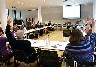 Das Bild zeigt eine Gruppe von Menschen, die an einem langen, U-förmigen Tisch in einem hellen Konferenzraum sitzen. Viele Teilnehmer haben ihre Hände zu einer Abstimmung erhoben. Auf dem Tisch befinden sich Bücher, Papiere, Getränke und Laptops. Im Hintergrund ist eine Leinwand mit einem Projektor zu sehen. Der Raum ist modern und hell eingerichtet.