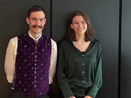 Zwei lächelnde Personen vor dunkler Wand, links ein Mann mit Schnurrbart und lila Weste, rechts eine Frau mit Brille und grünem Hemd.