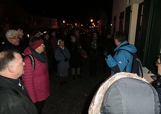 Das Bild zeigt eine Gruppe von Menschen bei Nacht, die an einem Gebäude stehen und aufmerksam einem Mann zuhören, der etwas erklärt. Der Mann trägt eine blaue Jacke und einen Rucksack. Die Gruppe steht auf einem gepflasterten Gehweg vor einem Gebäude mit Fenstern. Im Hintergrund sind weitere Gebäude und Straßenbeleuchtung zu sehen.