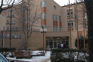 Das Bild zeigt das Don Bosco Haus in Wien. Es ist ein mehrstöckiges Gebäude in hellen Farben mit mehreren Fenstern und einem modernen Eingangsbereich. Vor dem Gebäude befinden sich Büsche, ein beleuchteter Weg und ein Schild mit dem Namen des Hauses. Im Vordergrund ist ein Teil eines Autos und eine Holzkonstruktion mit Figuren zu sehen. Der Schnee auf dem Boden deutet auf die Jahreszeit Winter hin.