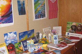Das Bild zeigt einen Tisch mit verschiedenen religiösen Broschüren, Karten, Büchern und Kerzen. An der Wand dahinter hängen Kalender und weitere religiöse Bilder. Die Materialien sind vielfältig und scheinen verschiedene Altersgruppen anzusprechen. Ein Kalender mit dem Jahr 2010 ist sichtbar.