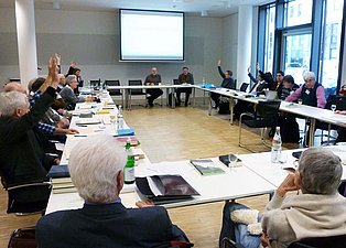 Das Bild zeigt eine Gruppe von Menschen, die an einem großen, U-förmigen Tisch in einem hellen Konferenzraum sitzen. Viele Teilnehmer haben für eine Abstimmung ihre Hände gehoben. Auf dem Tisch liegen Bücher, Papiere und Getränke. Im Hintergrund ist eine Leinwand mit einer Präsentation zu sehen. Der Raum ist modern und hell eingerichtet.