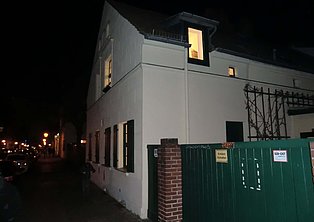 Das Bild zeigt ein weißes einstöckiges Gebäude bei Nacht. Ein Teil des Gebäudes ist beleuchtet, während der Rest im Dunkeln liegt. Ein grüner Zaun mit einem kleinen Tor ist im Vordergrund zu sehen. Im Hintergrund sind Straßenbeleuchtung und Autos zu erkennen. Das Bild wirkt ruhig und etwas geheimnisvoll.