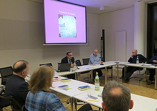 Das Bild zeigt eine Konferenz. Mehrere Personen sitzen an einem U-förmigen Tisch. Eine Person spricht und präsentiert eine Folie mit dem Titel "To Synthesize the Hopes" auf einer Leinwand. Auf dem Tisch stehen Wasserflaschen und liegen Dokumente. Der Raum ist hell und modern eingerichtet.