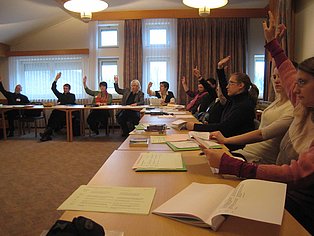 Das Bild zeigt eine Gruppe von Menschen, die an einem langen Tisch in einem Konferenzraum sitzen. Viele von ihnen haben ihre Hände zu einer Abstimmung erhoben. Auf dem Tisch liegen Papiere und Bücher. Der Raum ist hell und freundlich eingerichtet. Das Bild vermittelt den Eindruck einer aktiven und interaktiven Gruppensitzung.