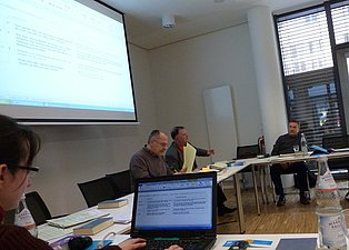 Das Bild zeigt eine Konferenz in einem modernen Raum. Mehrere Personen sitzen an einem langen Tisch, auf dem Laptops, Papiere und Getränke stehen. An der Wand ist eine Leinwand mit einer Präsentation zu sehen. Eine Person im Vordergrund ist an einem Laptop beschäftigt. Die Atmosphäre wirkt konzentriert und arbeitsorientiert.