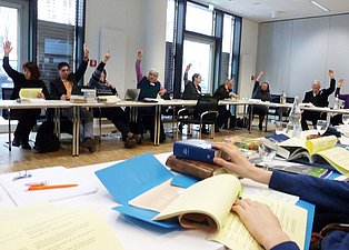 Das Bild zeigt eine Gruppe von Menschen, die an einem langen Tisch sitzen und ihre Hände heben. Sie nehmen an einer Konferenz teil, bei der gerade eine Abstimmungen erfolgt. Auf dem Tisch liegen Bücher, Papiere und Getränke. Der Raum ist hell und modern eingerichtet.