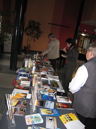 Das Bild zeigt einen langen Tisch, auf dem eine Vielzahl von Büchern, Broschüren und anderen Druckerzeugnissen ausgestellt sind. Zwei Personen stehen am Tisch, eine scheint etwas zu betrachten, die andere isst etwas. Im Hintergrund ist ein Raum mit Pflanzen und weiteren Personen zu sehen. 