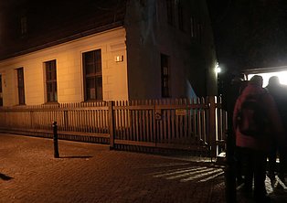 Das Bild zeigt eine Gruppe von Menschen, die nachts vor einem hell erleuchteten Gebäude stehen. Das Gebäude ist hellbeige und hat einen kleinen, weißen Holzzaun davor. Der Gehweg ist gepflastert. Die Atmosphäre ist ruhig und dunkel.