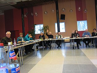Das Bild zeigt eine Gruppe von Menschen, die an einem langen, U-förmigen Tisch in einem großen Raum sitzen. Mehrere Personen haben ihre Hände erhoben. Der Raum hat rote und beigefarbene Wände und große Fenster. Auf dem Tisch liegen Papiere und Getränke. 