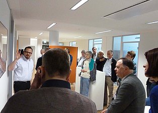 Das Bild zeigt eine Gruppe von Menschen in einem hellen, modernen Raum. Eine Person steht im Vordergrund vor einem nicht zu erkennenden Bild und erläutert die Darstellung. Die Atmosphäre wirkt informell und diskussionsorientiert. Im Hintergrund sind weitere Personen zu sehen.