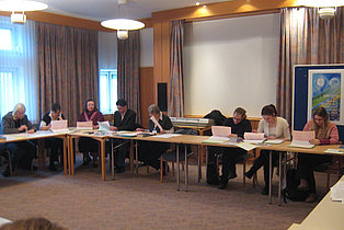 Das Bild zeigt eine Gruppe von etwa acht Personen, die um einen langen Tisch in einem Konferenzraum sitzen. Sie nehmen an einer Besprechung teil und lesen oder schreiben an Papieren. Der Raum ist hell und freundlich eingerichtet mit Vorhängen, einem Projektor und einem Keyboard im Hintergrund. Die Atmosphäre wirkt konzentriert und arbeitsam.