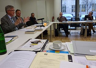 Das Bild zeigt eine Gruppe von Personen, die an einem langen, U-förmigen Tisch in einem hellen Konferenzraum sitzen. Eine Person gestikuliert, während sie spricht. Die anderen Teilnehmer sitzen und hören zu. Auf dem Tisch befinden sich Getränke, Papiere und Bücher. Im Hintergrund sind Fenster und ein Teil der Straße zu sehen. Der Raum wirkt modern und professionell.