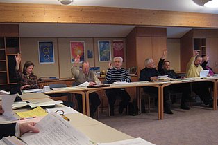Das Bild zeigt eine Gruppe von etwa acht Personen, die um einen langen Tisch in einem hellen Raum sitzen. Mehrere Personen haben ihre Hände zu einer Abstimmung erhoben. Auf dem Tisch liegen Papiere und Bücher. Die Wände sind mit Holz verkleidet und mit Bildern und Postern dekoriert. Der Raum wirkt freundlich und informell, die Atmosphäre ist aktiv und interaktiv.