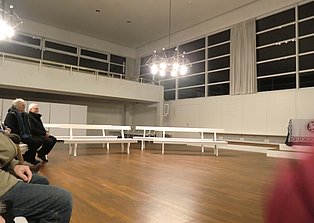 Das Bild zeigt einen großen, hellen Raum mit einem Holzboden. Im Raum stehen mehrere weiße Bänke in Reihen angeordnet. An einem Ende des Raumes befindet sich eine erhöhte Plattform mit einem Altar. Zwei Personen sitzen auf einer Bank im Vordergrund. Die Wände sind weiß und große Fenster lassen Licht in den Raum. Der Raum wirkt ruhig und geräumig.