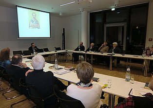 Das Bild zeigt eine Gruppe von Menschen, die an einem langen, U-förmigen Tisch in einem Konferenzraum sitzen. Ein Mann sitzt an einem Ende des Tisches und hält eine Präsentation. Die anderen Teilnehmer sitzen und hören zu. Auf dem Tisch stehen Getränke und es liegen Papiere bereit. Der Raum ist gut beleuchtet und modern eingerichtet.