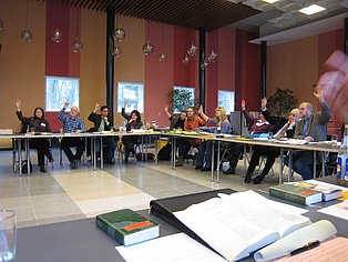 Das Bild zeigt eine Gruppe von Menschen, die um mehrere Tische in einem großen Raum sitzen. Viele von ihnen haben ihre Hände erhoben für eine Abstimmung erhoben. Der Raum ist modern eingerichtet mit roten und beigefarbenen Wänden und großen Fenstern. Auf den Tischen liegen Papiere und Bücher. Die Atmosphäre wirkt aktiv und interaktiv.