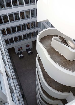 Das Bild zeigt eine moderne, geschwungene Treppe in einem hellen Atrium. Die Treppe windet sich spiralförmig nach oben und ist aus weißem Material mit braunen Marmorstufen gefertigt. Im Hintergrund ist ein Innenhof mit weiteren Büros zu sehen. Das Gesamtbild wirkt modern und architektonisch ansprechend.