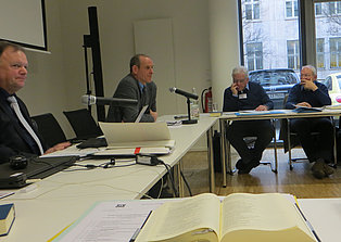 Das Bild zeigt eine Konferenzszene mit vier Männern, die an einem langen U-förmigen Tisch sitzen. Ein Mann im Vordergrund spricht, während die anderen zuhören. Auf dem Tisch liegen Dokumente, Mikrofone und Getränke. Im Hintergrund ist ein Fenster mit Blick auf eine Straße und geparkte Autos zu sehen. Der Raum ist hell und modern eingerichtet.