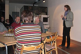 Das Bild zeigt eine Frau, die vor einer kleinen Gruppe von Zuhörern steht und spricht. Sie trägt einen grauen Blazer und dunkle Hose. Die Zuhörenden sitzen an Tischen und scheinen aufmerksam zu sein. Im Hintergrund sind weitere Personen und ein Bücherregal zu erkennen. Der Raum wirkt informell und gemütlich.